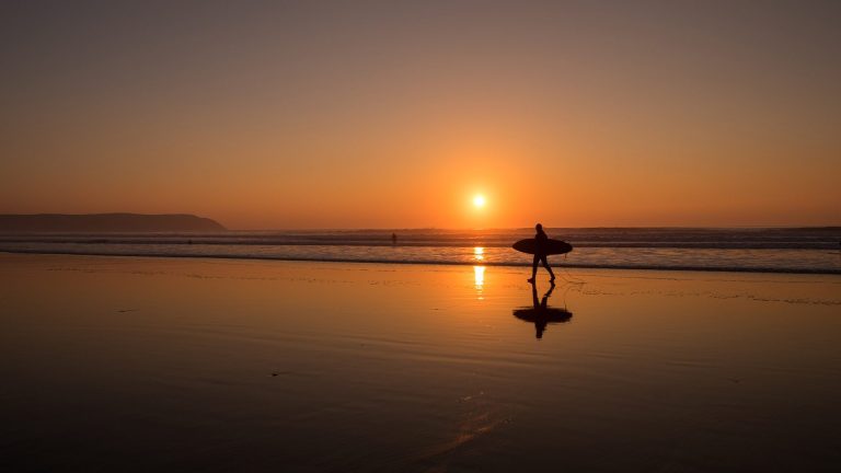 A person carrying a surfboard along the beach at sunset