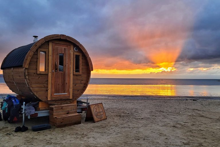 Barrel sauna on the beach at Oxwich Bay with the sun setting over the sea